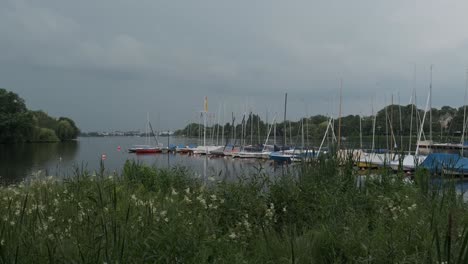 Streak-of-forked-lightning-over-the-city-of-Hamburg-shrouded-in-grey-cloud-as-seen-from-the-Outer-Alster-lake-with-moored-boats-at-a-jetty-over-greenery-in-the-foreground