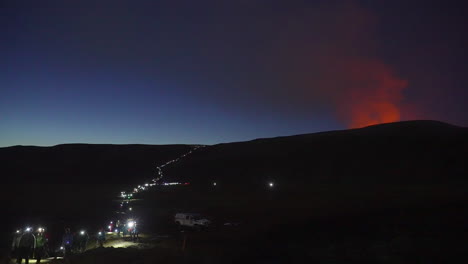 Crowd-of-people-with-forehead-lamps-hiking-back-at-blue-hour-after-visiting-eruption-of-volcano-in-background