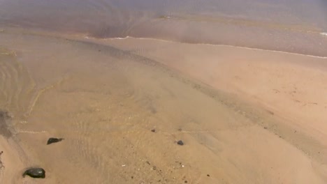 Slowly-panning-from-looking-down-at-a-sandy-beach-out-to-deeper-water-and-a-paddleboarder