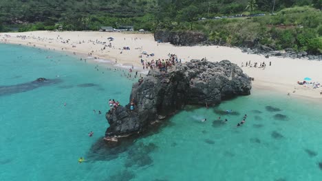 Drone-flying-towards-people-having-fun,-jumping-from-rocks-on-Pristine-Sand-beach,-Hawaii