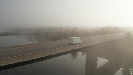 Commercial-transportation,-tractor-trailer-and-box-truck-crossing-highway-bridge-with-blanket-of-fog-hanging-over-river-and-landscape