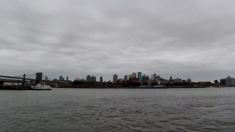 Ein-Zeitraffer-über-Dem-East-River-Der-Industriellen-Skyline-Von-Brooklyn-Aus-Manhattan,-Während-An-Einem-Bewölkten-Tag-Boote-Vorbeifahren