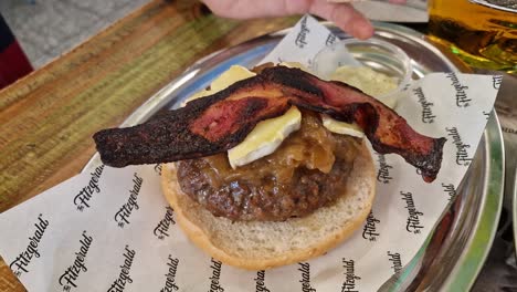 Mano-De-Una-Persona-Levantando-El-Pan-De-Una-Hamburguesa-Gourmet-Con-Queso-De-Cabra,-Cebolla-Caramelizada-Y-Tocino-Tostado-En-Una-Bandeja