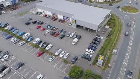 Drone-rising-establishing-shot-of-a-Lidl-supermarket-in-Bude,-Cornwall,-United-Kingdom,-in-July-2024,-showing-the-layout-of-the-parking-area,-nearby-streets,-and-adjacent-shops