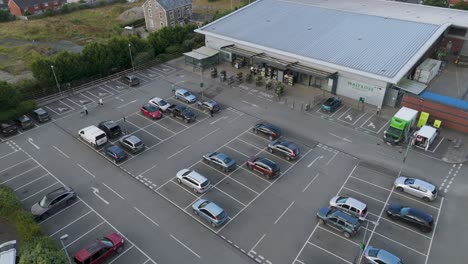 Aerial-fly-out-view-of-Waitrose-supermarket-and-its-carpark,-providing-a-comprehensive-view-of-the-retail-space-and-its-location,-Holsworthy,-Devon,-United-Kingdom,-June-2024