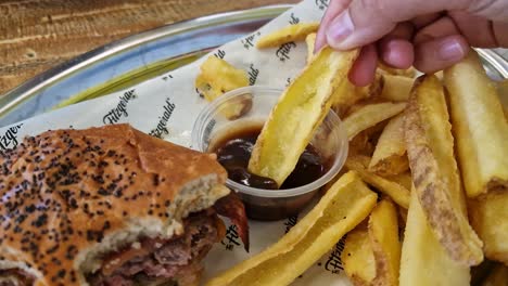 Hand-of-a-person-dipping-a-chip-in-barbecue-sauce-next-to-a-hamburger-on-a-tray