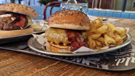 Cheeseburger-with-bacon-and-fries-on-a-plate,-on-a-table-with-another-burger-in-the-background