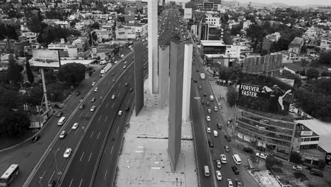 Black-and-white-drone-shot-of-the-"Torres-de-Satélite",-in-the-suburbs-of-Mexico-City,-a-cloudy-afternoon-with-flowing-traffic