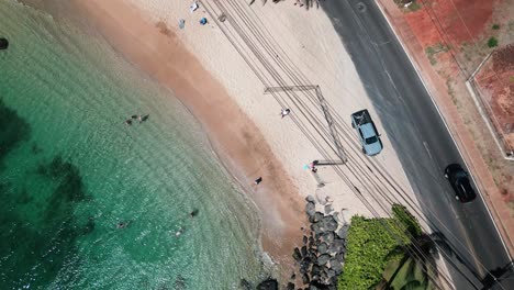 Drone-view-of-shallow-water-beach-with-people,-truck,-clear-water,-and-powerlines