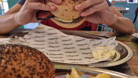 A-person-holds-a-large-hamburger-with-a-black-bread-and-bacon,-fries-and-sauce-on-a-tray-on-a-table-with-branded-paper