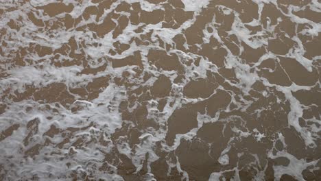 Aerial-view-of-gentle-waves-creating-frothy-patterns-as-they-wash-onto-a-sandy-beach