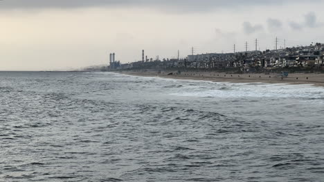 Olas-Ondulantes-Hacia-La-Mansión-Multimillonaria-Bordeada-De-La-Costa-De-La-Playa-De-Manhattan-Con-Una-Planta-De-Energía-De-Vapor-Visible-En-La-Distancia-En-Una-Nube-Y-Un-Día-Sombrío