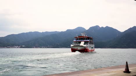 Das-Rot-weiße-Jr-Fährschiff-Fährt-An-Einem-Kühlen,-Bewölkten-Tag-In-Japan-Von-Der-Uferpromenade-über-Das-Wasser-In-Richtung-Der-Insel-Itsukushima