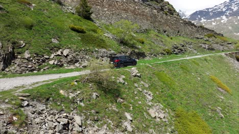 Black-car-drives-slowly-up-dirt-trail-in-scenic-alpine-landscape-with-snowy-mountains