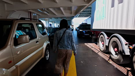 Woman-with-jeans-jacket-and-sun-glasses-is-walking-in-slow-motion-between-the-cars,-trucks-on-a-ferry,-back-view