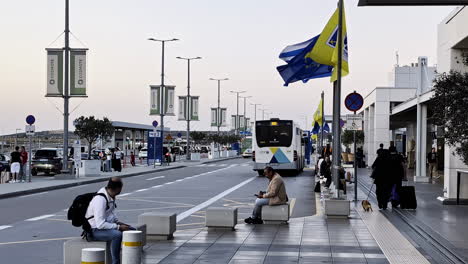 Statische-Aufnahme-Des-Außenbereichs-Für-Busse-Und-Taxis-Des-Flughafens,-Ankunft-Und-Abflug,-Athen,-Griechenland,-Fahnen-Bewegen-Sich-Im-Wind,-Menschen-Sitzen-Und-Warten