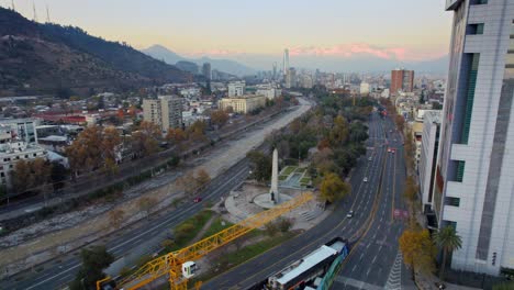 Aerial-orbit-establishing-of-the-Balmaceda-Obelisk-in-Balmaceda-Park-with-the-Mapocho-river-in-Santiago-de-Chile,-sunset-with-the-Andes-mountain-range