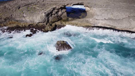 aerial-view-of-a-dramatic-rock-arch-jutting-out-over-the-ocean,-with-powerful-waves-crashing