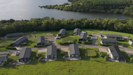 Vista-Aérea-De-Casas-De-Vacaciones-De-Lujo-Junto-A-Un-Lago-Sereno-Durante-La-Hora-Dorada,-Ubicadas-En-La-Campiña-De-Devon