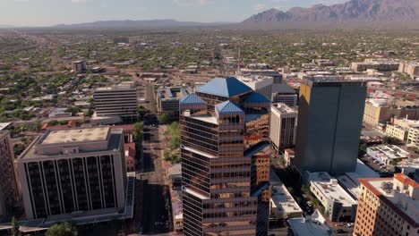 Aerial-Shot-of-Shiny-Rose-Gold-Building-in-Downtown-Tucson,-Arizona-with-Mountains-on-Horizon-and-Vast-Urban-Landscape