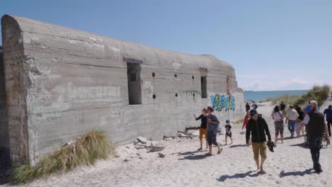 Toma-Panorámica-Manual-De-Turistas-Caminando-Desde-Y-Hacia-El-Lugar-Donde-Se-Encuentran-El-Mar-Báltico-Y-El-Mar-Del-Norte,-Pasando-Por-Un-Antiguo-Búnker-De-La-Segunda-Guerra-Mundial-En-La-Playa-De-Grenen