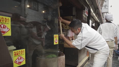 A-staff-member,-along-with-Mitsuo-Nakatani,-pounds-a-batch-of-mochi-with-a-wooden-mallet-as-part-of-the-traditional-mochitsuki-pounding-method-at-Nakatanidou-Mochi-Shop-in-Nara,-Japan,-medium-track