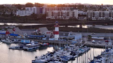 Aerial-footage-of-Oceanside-Harbor-at-sunrise,-showing-the-lighthouse,-colorful-waterfront-buildings,-and-boats-docked-along-harbor-with-the-cityscape-in-the-background