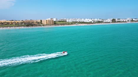 A-person-is-on-a-jet-ski,-speeding-across-the-water-near-a-beach-with-many-buildings-and-houses-in-the-background-at-Mahdia