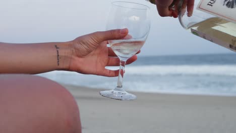 Person-pours-glass-of-wine-while-sitting-on-beach-looking-out-to-ocean-waves