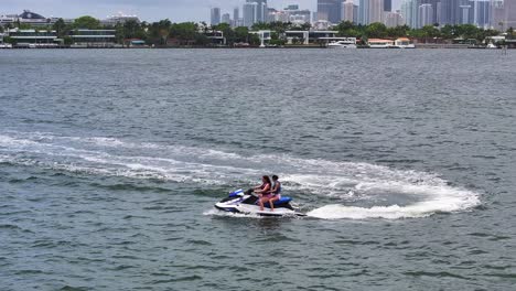 Zwei-Mädchen-Auf-Jetski-Auf-Dem-Fluss-Vor-Der-Venezianischen-Insel-Und-Dem-Wolkenkratzer-In-Der-Innenstadt-Im-Hintergrund