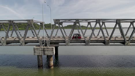 Rotes-Tuk-Tuk-Fährt-An-Einem-Sonnigen-Tag-über-Eine-Metallbrücke-über-Einem-Ruhigen-Fluss