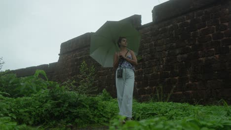 A-woman-holding-an-umbrella-stands-by-an-ancient-stone-wall-on-a-rainy-day