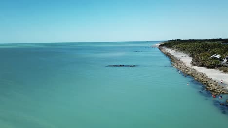 Aerial-view-of-a-rocky-shoreline-extending-into-a-calm-blue-sea,-with-beachgoers-and-a-wooden-walkway