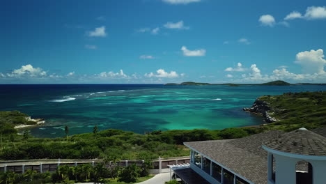 Drone-flying-low-over-buildings,-toward-turquoise-sea,-sunny-day-in-Antigua