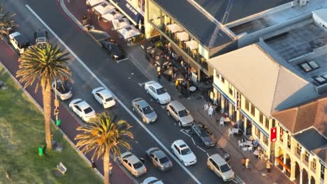 Primer-Plano-De-Un-Dron-De-Un-Paseo-Marítimo-En-La-Playa-De-Camps-Bay-En-Ciudad-Del-Cabo,-Sudáfrica:-Coches-Circulando-Por-La-Calle-Junto-A-Tiendas-Y-Gente-Local;-Hay-Palmeras-Al-Costado-De-La-Carretera