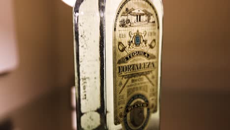 Botella-De-750-Ml-De-Tequila-Blanco-Fortaleza-Colocada-Sobre-Una-Mesa-De-Vidrio,-Reflejo-Iluminado-Por-Una-Lámpara-Cálida-Que-Resalta-La-Etiqueta-Antigua,-Cámara-Panorámica-Hacia-Arriba-Y-Hacia-Abajo