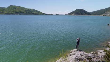 Young-male-photographer-hiker-standing-on-a-rock-at-the-edge-of-a-mountain-lake-photographing-Spring-Sunny-day