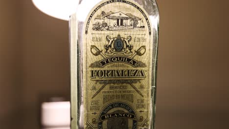 Toma-Estática-De-Una-Botella-De-750-Ml-De-Tequila-Fortaleza-Blanco-Y-La-Etiqueta-Frontal-Nom-1493-Botella-Guillermo-Sauza-Iluminada-Desde-Atrás-Por-Una-Lámpara-Cálida-Ambiente-Oscuro