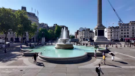 Scenic-view-of-people-enjoying-a-sunny-morning-at-Trafalgar-Square-fountain-in-London,-with-clear-blue-skies-and-historical-architecture