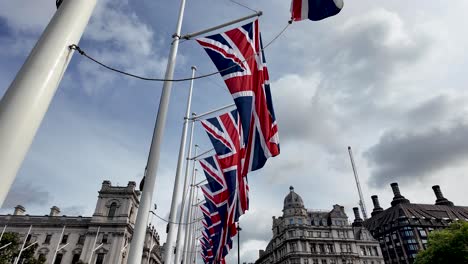 A-series-of-Union-Jack-flags-flutter-in-the-breeze-against-a-backdrop-of-historic-architectural-buildings-and-an-overcast-sky-in-London,-symbolizing-British-pride-and-heritage