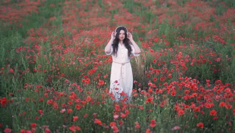 general-plan-beautiful-young-dark-haired-woman-with-long-hair-putting-on-wireless-headphones,-starting-to-dance-and-sing-in-a-dress-against-a-background-of-wildflowers-and-red-poppies