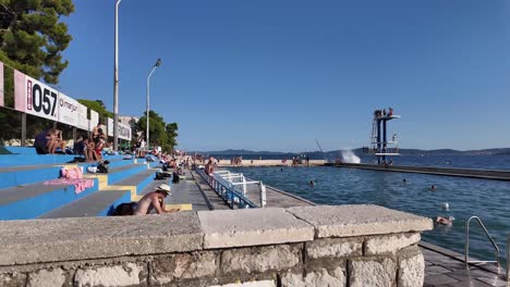 Kolovare-Strand-Mit-Meeresbecken-Und-Sprungbrett-Und-Menschen-Beim-Sonnenbaden