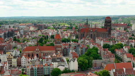 Aerial-view-of-Gdańsk's-old-town,-showcasing-historic-buildings-with-red-tiled-roofs,-churches,-and-green-spaces-against-a-backdrop-of-a-vast-landscape