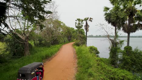 Tuk-tuk-driving-on-a-dirt-road-by-a-lush-riverbank-surrounded-by-tropical-greenery