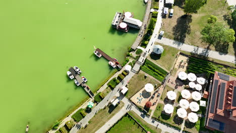 Aerial-perspective-of-a-beautiful-lakeside-town-in-Warmia-Masuria,-featuring-green-lake,-docks,-boats,-and-vibrant-buildings-on-a-sunny-day,-Poland