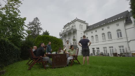 Time-lapse-of-a-group-of-people-sitting-and-standing-outside-in-the-garden-on-a-table,-talking-on-a-cloudy-day-with-a-beautiful-villa-in-the-background,-static,-copy-space