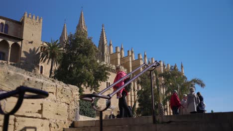 Tourists-walking-around-the-Cathedral-of-Mallorca,-appreciating-the-historical-architecture-and-enjoying-the-clear,-sunny-weather