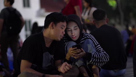 Asian-Couple-Gazing-Down-At-A-Smartphone-Screen-In-Taman-Fatahilla-Square,-Jakarta,-Indonesia