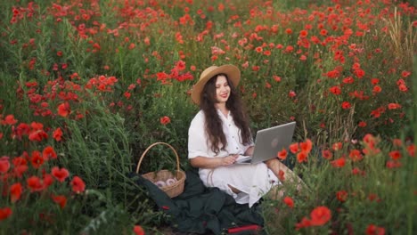 cheerful-dark-haired-girl-in-a-straw-hat-sits-in-a-field-of-wildflowers-and-red-poppies,-smiling-dreamily-at-a-joyful-message-on-her-laptop