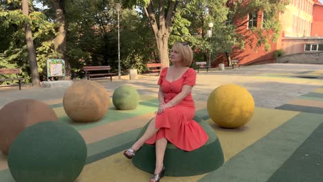 Pull-out-on-a-blonde-woman-in-an-orange-dress-sitting-on-a-half-play-rubber-ball-on-a-playground-and-enjoying-her-time